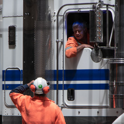A worker wearing protective headphones looks up towards the cab of a commercial truck. The driver leans out the window to see the worker wearing headphones