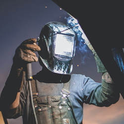 A welder wearing a protective face mask while using a welding gun on a metal joint. 